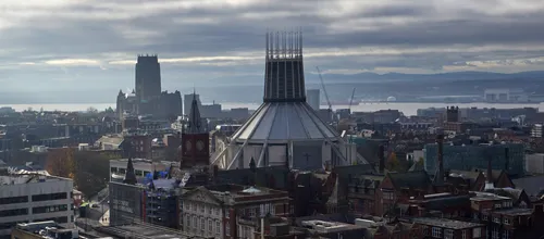 This urban landscape photograph shows the city of Liverpool in the United Kingdom during a late autumn morning. The main subject is the impressive pair of cathedrals; in the foreground stands the Liverpool Metropolitan Cathedral, a modern church known for its unique circular design and prominent spire, while in the distance the traditional Liverpool Cathedral rises above the city with its tall, historic tower. The morning light, softened by clouds, highlights the architectural contrasts between these significant religious landmarks. The image captures the density of Liverpool’s cityscape, with a mixture of historic and contemporary buildings surrounding the cathedrals. The River Mersey and distant hills are visible in the background, providing a scenic backdrop to the urban scene and emphasizing Liverpool’s status as a prominent city in the United Kingdom.